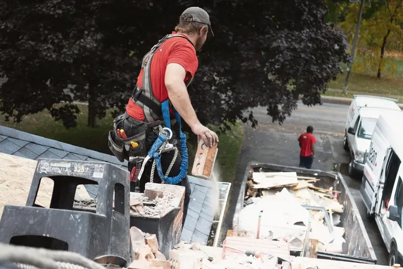 A tradesman on a roof dropping old bricks into a waste container